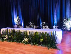 the table is set up with white chairs and green plants on it, along with blue drapes