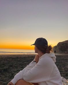 a woman sitting on top of a sandy beach next to the ocean at sun set