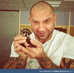 a bald man holding a baby raccoon in his right hand and smiling at the camera