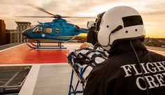 a man wearing a white helmet and black jacket standing next to a helicopter on top of a building