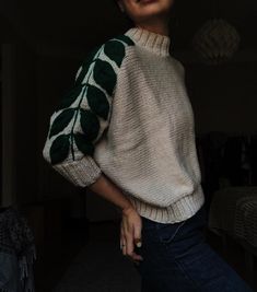 a woman standing in front of a bed wearing jeans and a sweater with leaves on it