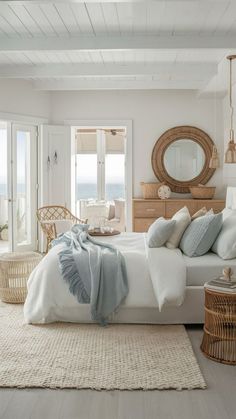 a bedroom with white walls and flooring has a large bed, wicker chair, rattan side tables, and an oval mirror on the wall