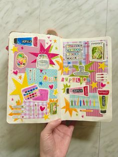 a hand holding an open notebook with various stickers on the pages and in front of it