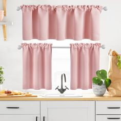 a kitchen window with pink curtains and white cabinets in front of the window is a potted plant