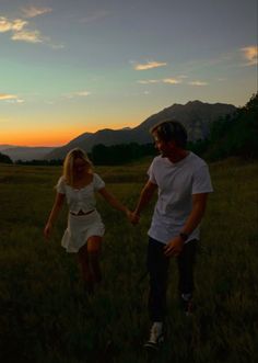 a man and woman holding hands while walking through a field at sunset with mountains in the background