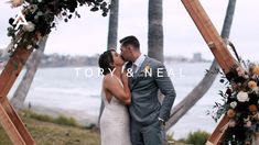 a newly married couple kissing under an arch with flowers and greenery in front of the ocean