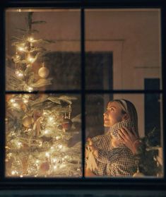 a woman standing in front of a window next to a christmas tree with lights on it