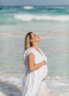 a pregnant woman standing in the ocean with her arms around her waist, looking up at the sky