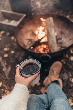 a person holding a coffee cup in front of a campfire with their feet on the ground