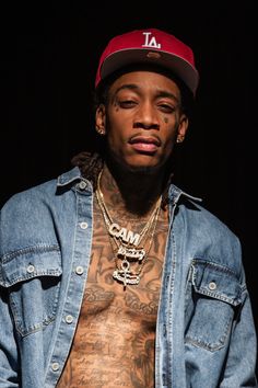 a man with tattoos wearing a denim jacket and red baseball cap is looking at the camera