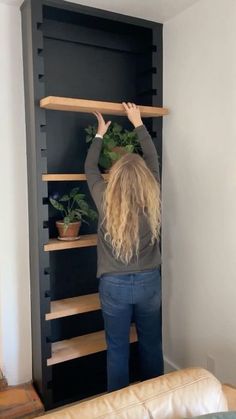 a woman standing in front of a black book shelf holding a plant on top of it