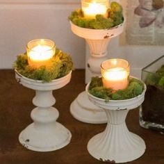 three white candle holders filled with moss and lit candles on top of a wooden table