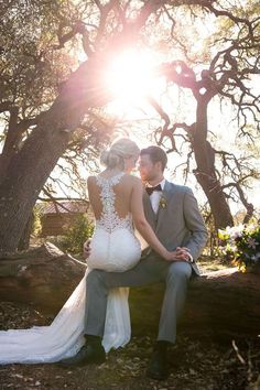 a bride and groom are sitting on a log in front of some trees with the sun behind them