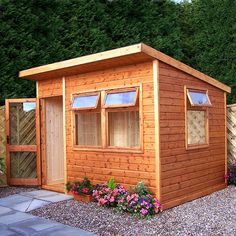 a small wooden shed with windows on the side and flowers in the yard around it