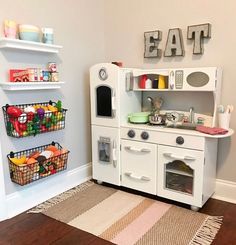 a white play kitchen with lots of food on the shelves and toys in baskets next to it