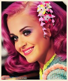 a woman with bright pink hair and flowers in her hair smiles at the camera while wearing a colorful outfit