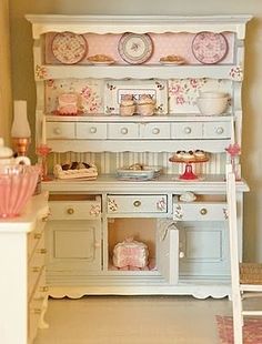 a doll house kitchen with white furniture and pink accessories on the shelves, along with other items