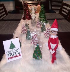 an elf is sitting on the table with christmas trees for sale in front of him