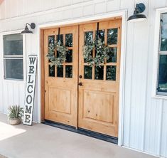 two wooden doors on the side of a white building with welcome sign and potted plant