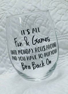 a wine glass with words on it sitting on a white sheet that says, it's all fun & games until monday rolls around and you have to put your bra back on