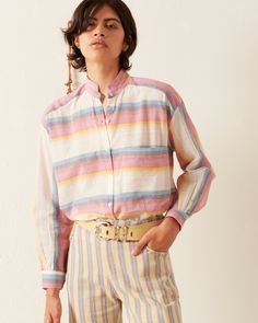 A person wearing the Kiki Sunset Shirt with Mother of Pearl buttons and matching striped pants stands against a plain background. They have short, dark hair and are posing with one hand in their pocket, showcasing a casual yet refined outfit that hints at artisanal craftsmanship. Sunset Shirt, Women Gathering, Shirt Cuff, Weaving Process, Hand Loom, Notched Collar, Mother Of Pearl Buttons, A Mother, Striped Shirt