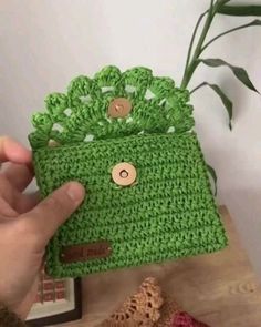 a hand holding a green crocheted purse next to a potted plant on top of a wooden table