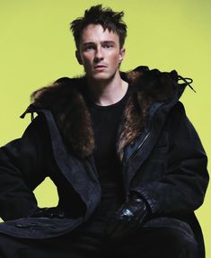 a man sitting on a stool wearing a black jacket and fur collared hoodie