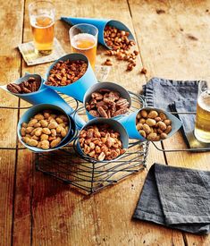 several bowls filled with nuts on top of a wooden table next to two glasses of beer