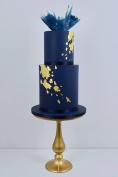 a three tiered blue and gold wedding cake with feathers on the top, against a white background