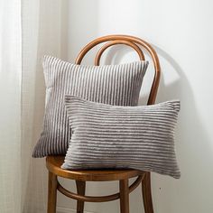 two gray pillows sitting on top of a wooden chair next to a white wall and window