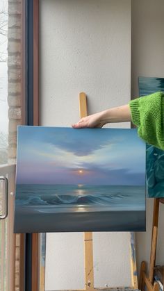 a person holding up a painting in front of a window with the sun going down