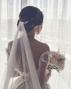 a woman wearing a wedding dress and holding a bouquet in front of a window with sheer curtains