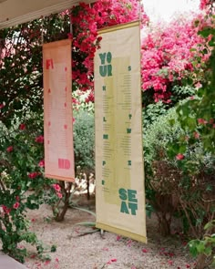 two banners are hanging from the side of a house in front of some pink flowers