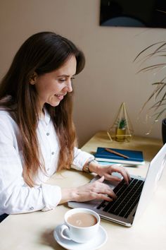 a woman sitting in front of a laptop computer on top of a desk next to a cup of coffee