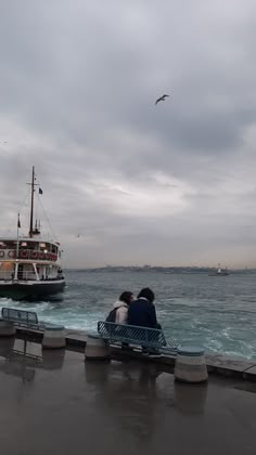 two people sitting on a bench looking out at the ocean with a boat in the background