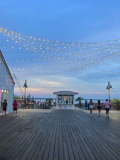 people are walking around on the boardwalk at dusk with string lights strung across the walkway