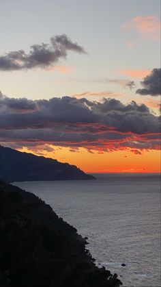 the sun is setting over the ocean with mountains in the backgrouund and clouds in the sky