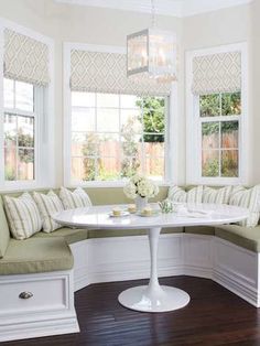 a corner table with white chairs and pillows on it in front of two large windows