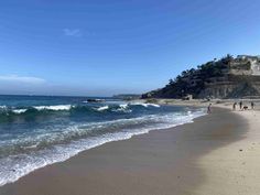 people are walking on the beach next to the ocean and cliffs in the distance, with waves crashing onto the shore