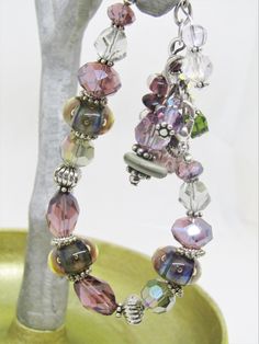 "This ARTISAN GLASS (Lampwork) BRACELET features 3 BOROSILICATE GLASS BEADS in shades of purple and green. The Boro glass beads are roughly 8x15mm and are spaced throughout the bracelet. There are also 10x14mm crystal tapered faceted ovals in purple and 8x12mm faceted crystal rondelles in the same purple; 5x8mm silver spacer beads, along with assorted clear and lavender faceted crystals. There are 7 assorted drops on the chain, inclding lampwork rondelles, Swarovski crystals, other glass, crysta Adjustable Glass Crystal Bracelet Gift, Silver Glass Beaded Bracelets For Gifts, Adjustable Glass Beaded Bracelets Nickel Free, Adjustable Silver Wearable Art Bracelet, Handmade Adjustable Glass Crystal Bracelet, Silver Glass Bracelets With Round Beads, Adjustable Nickel-free Jewelry In Wearable Art Style, Silver Adjustable Bracelets With Unique Variations, Adjustable Silver Bracelets With Unique Variations