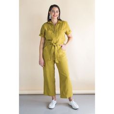 a woman standing in front of a white wall wearing a yellow jumpsuit and white sneakers