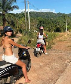 two women riding scooters on the side of a dirt road next to palm trees