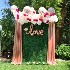 an arch decorated with balloons and the word love is displayed in front of a lush green wall