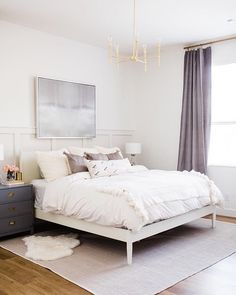 a white bed sitting in a bedroom next to a gray chest of drawers and a chandelier