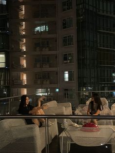 two women sitting on white couches in front of a tall building at night,