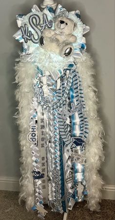 a white teddy bear with blue and silver decorations on it's chest, standing in front of a wall