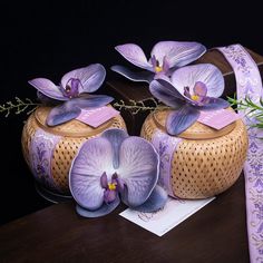 two purple flowers are sitting on top of a wooden box next to a ribbon and flower arrangement