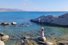 a woman standing on top of a rock covered beach next to the ocean with clear blue water