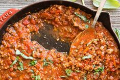 a skillet filled with meat and sauce on top of a wooden table next to a spoon