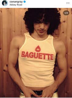 a man with curly hair wearing a white tank top and gold shorts is standing in front of a wooden wall